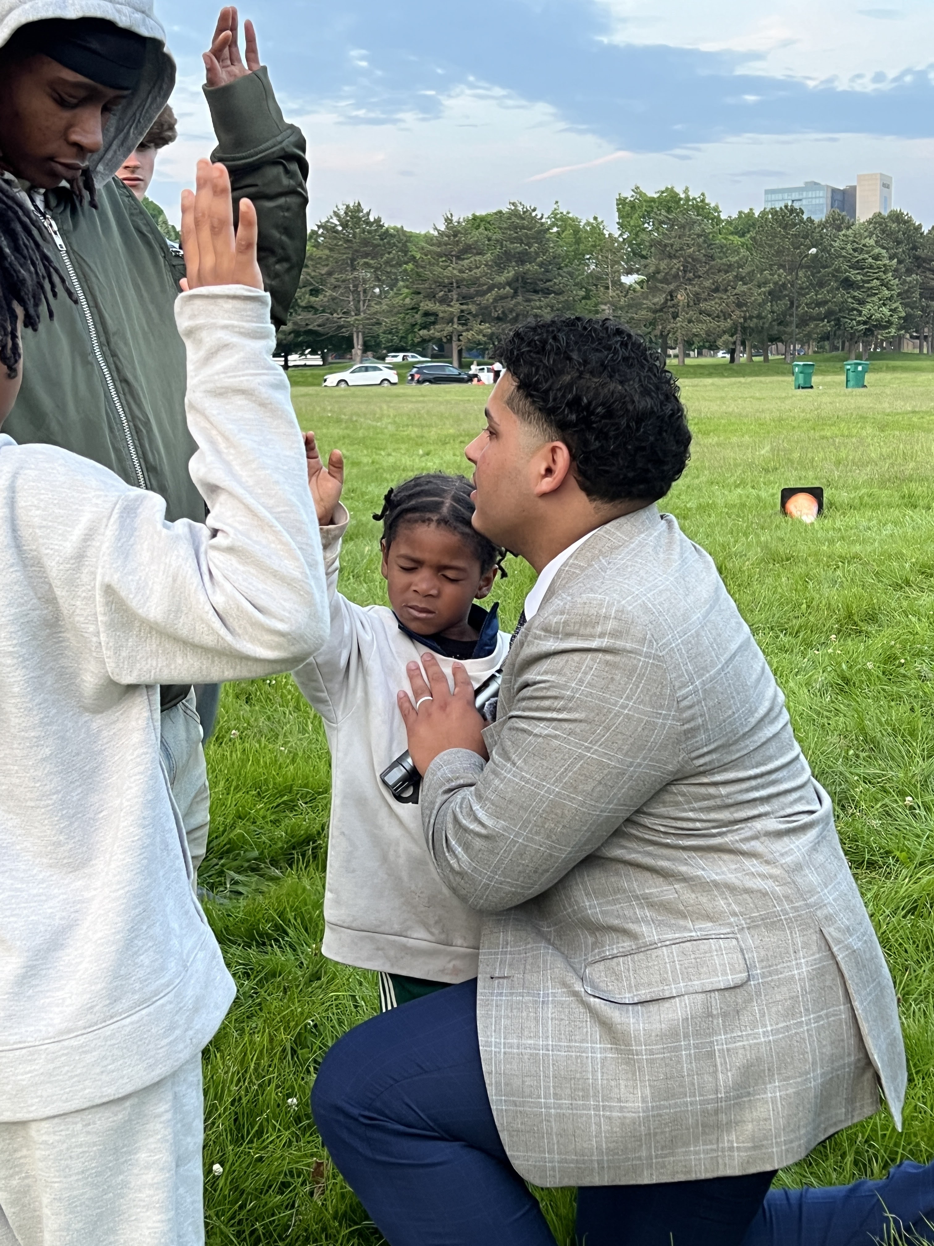 Izaiah kneeling in prayer with a young child outdoors at a Buffalo gathering