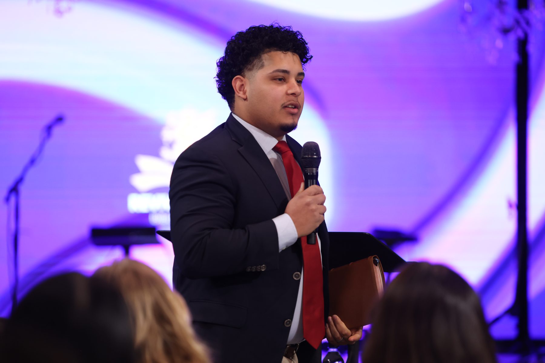 Evangelist Izaiah Diaz preaching on stage in a navy suit with microphone