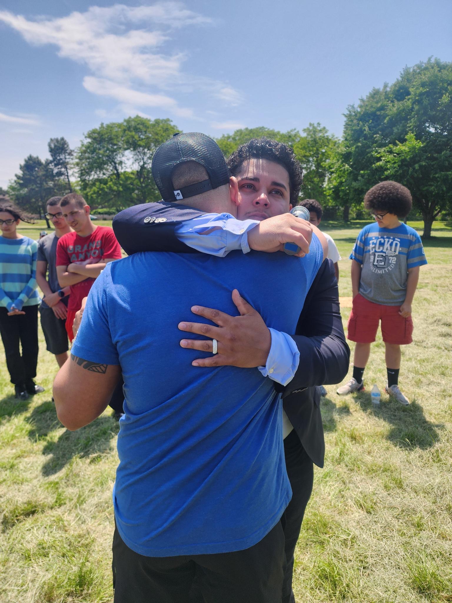 Evangelist Izaiah Diaz embracing a man in tears during outdoor ministry, surrounded by youth
